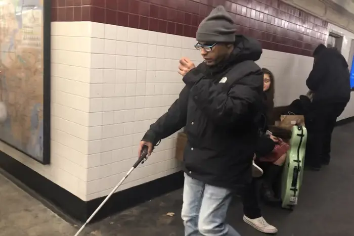 Bryan Velazquez walking on a subway platform, wearing a gray hat and glasses while holding a white cane in his right hand.